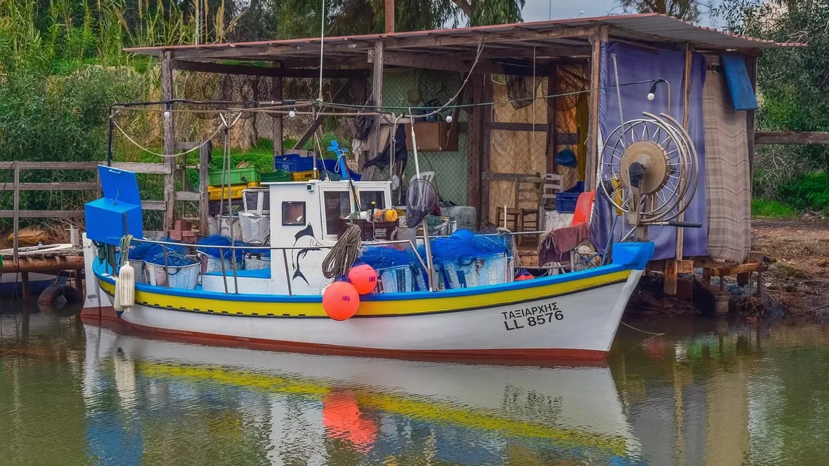 A white fishing boat with blue and yellow stripes, parked in front of a small wooden building on the shore of a body of water.