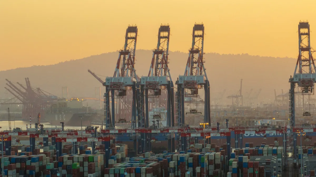 A large port with multiple shipping containers, cranes, and a mountain in the background.